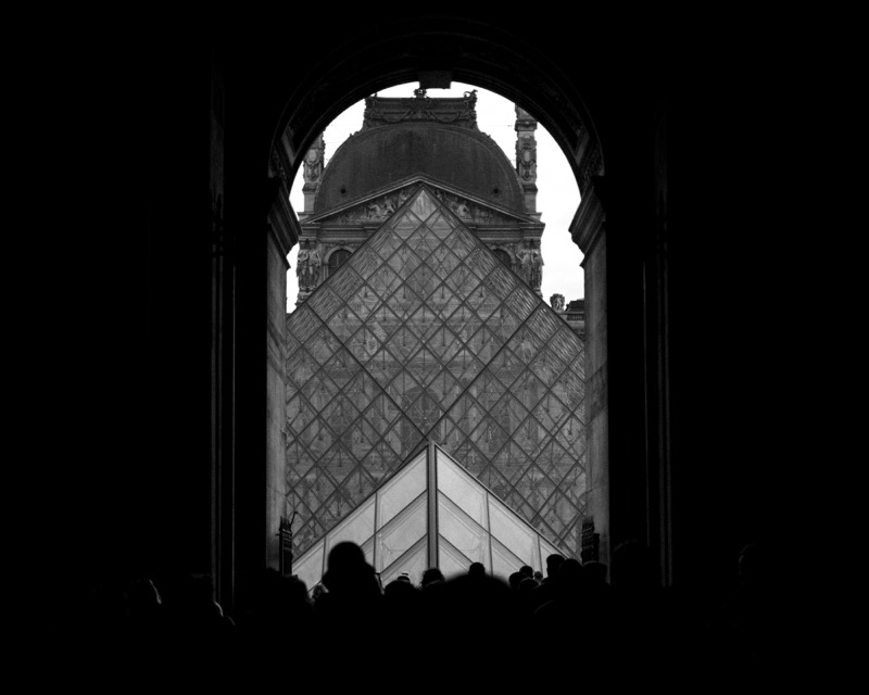 Louvre Pyramid seen through an archway, with silhouetted crowd in foreground.