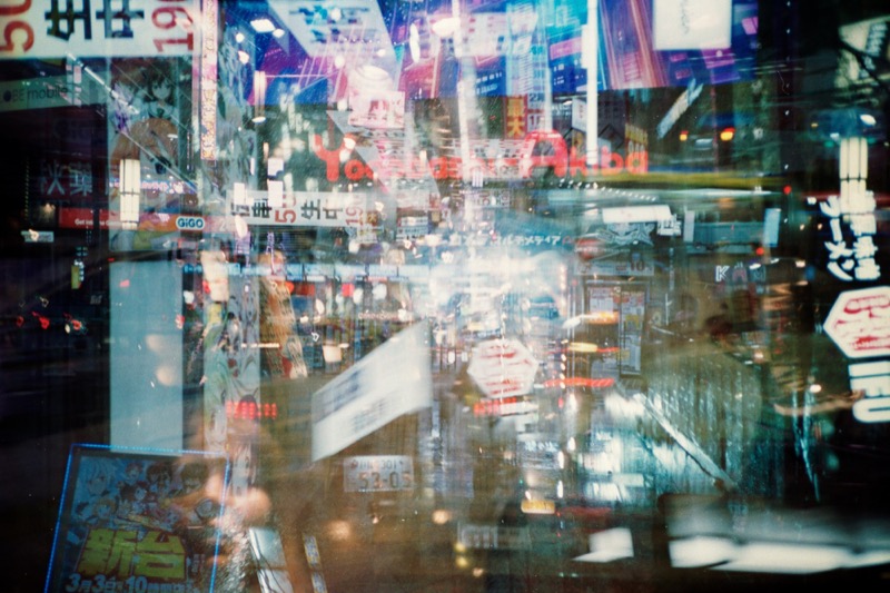 Double-exposed neon city street scene with signs, lights, and rain reflections.