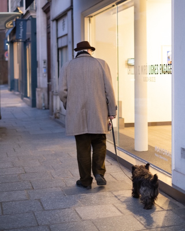 Man in a hat walks a small dog past a lit storefront at dusk.