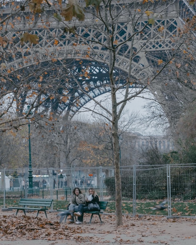Two people sit on a bench beneath the Eiffel Tower on a cold day.