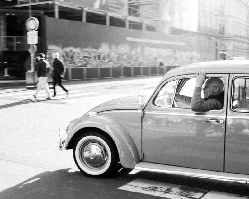 Driver in a vintage car shields his face while pedestrians cross nearby.