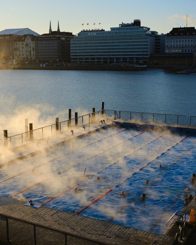 Steaming outdoor pool beside a harbor, with swimmers in winter light.