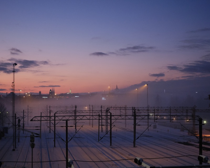 Snowy rail yard at twilight with fog, lights, and distant city skyline.