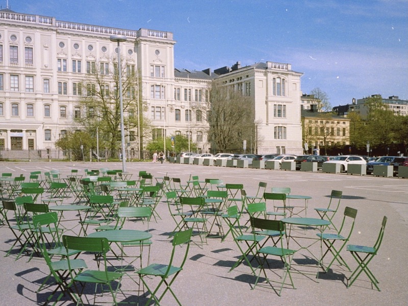 Empty green chairs and tables in a plaza in front of white buildings.