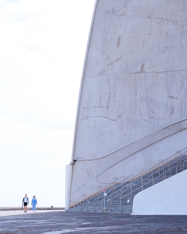 Two people walk beside massive concrete architecture and stairs under pale sky.
