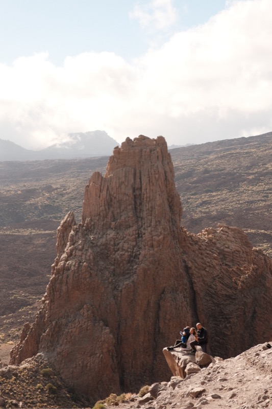 Hikers rest on boulders below a towering rock spire in hazy mountains.