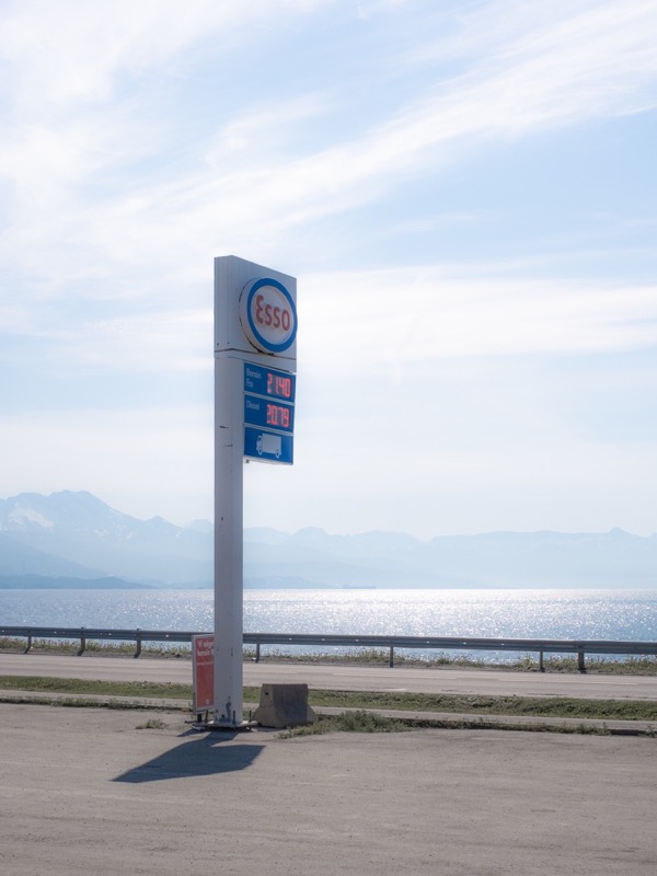 Esso price sign stands by a coastal road with mountains and sparkling water.