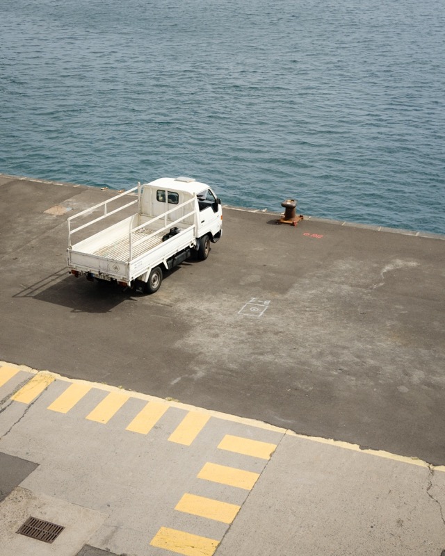 Small white truck parked on a harbor quay beside calm blue water.