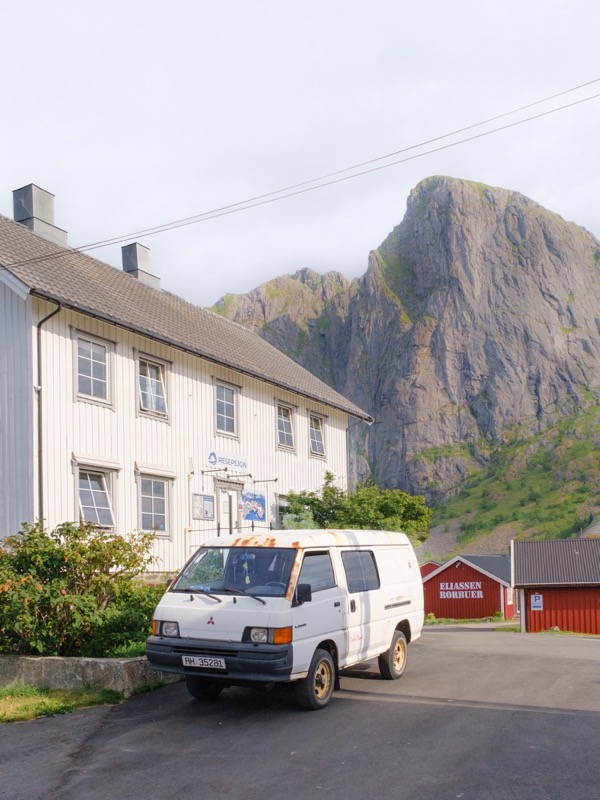 Old white van parked by a lodge, with steep mountain cliffs behind.