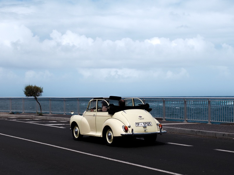 Cream convertible drives along a seaside road under a wide cloudy sky.