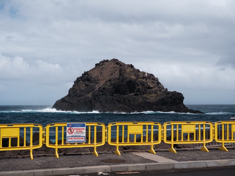 Rocky islet rises from rough sea behind yellow barriers and warning sign.