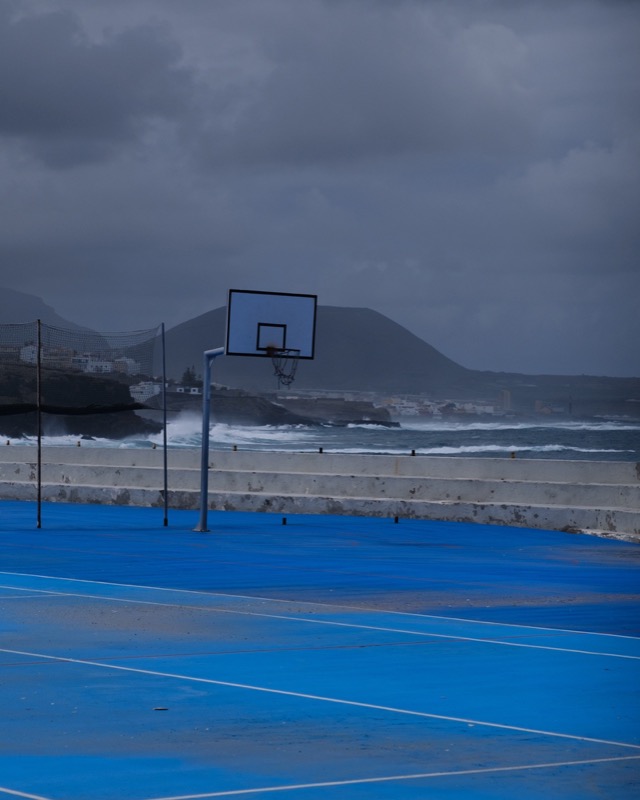 Blue outdoor basketball court sits beside crashing waves under dark storm clouds.