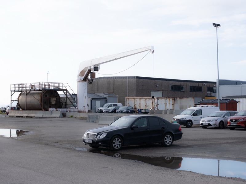 Black sedan parked in an industrial yard with a crane and warehouses.