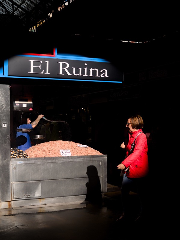 Woman in red jacket stands by the 'El Ruina' seafood stall in sharp sunlight.