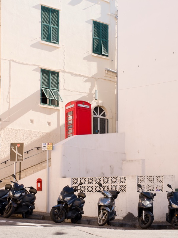 Red telephone booth sits on a white street above parked scooters.