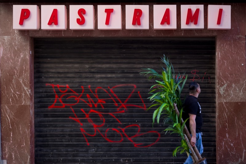 Man carries a tall plant past a shuttered shop beneath a 'Pastrami' sign.