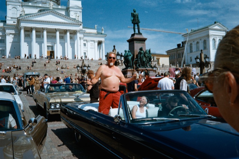Shirtless man stands in a convertible amid a crowded square and cathedral steps.