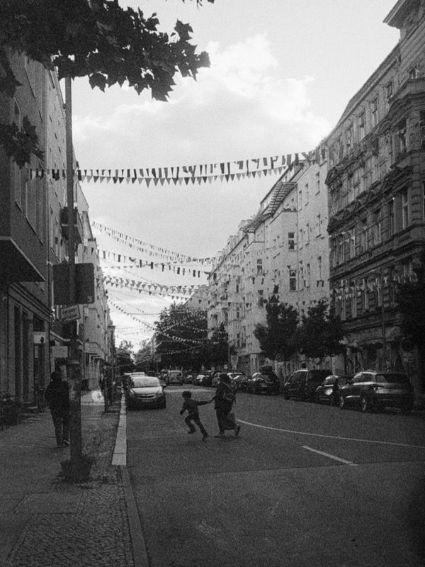 Black-and-white street scene with bunting overhead and children crossing the road.