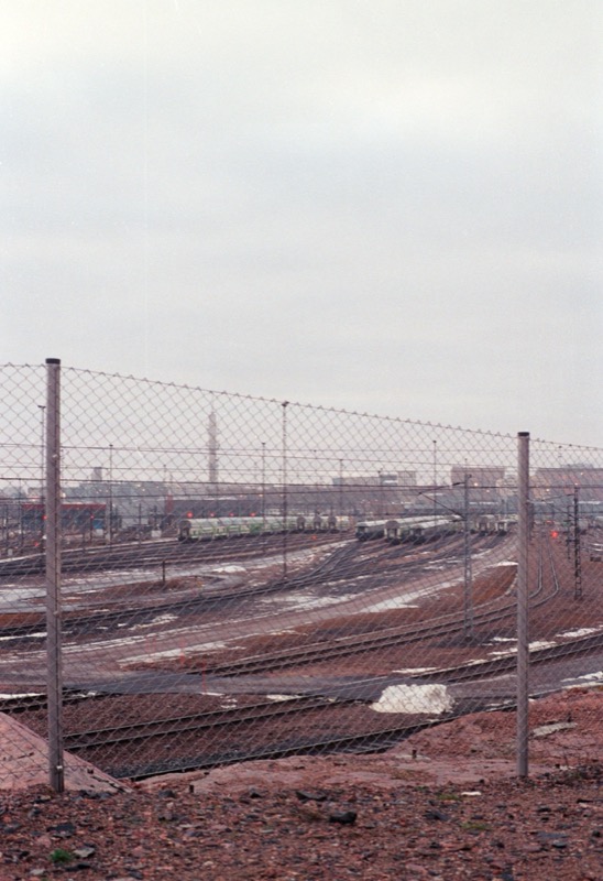 Rail yard seen through a chain-link fence under a pale, overcast sky.