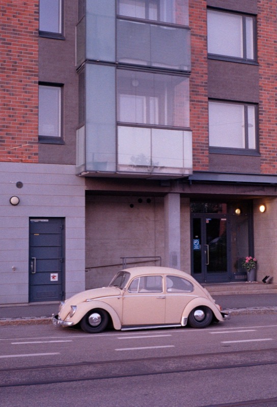 A beige Volkswagen Beetle is parked in front of a modern brick apartment building.