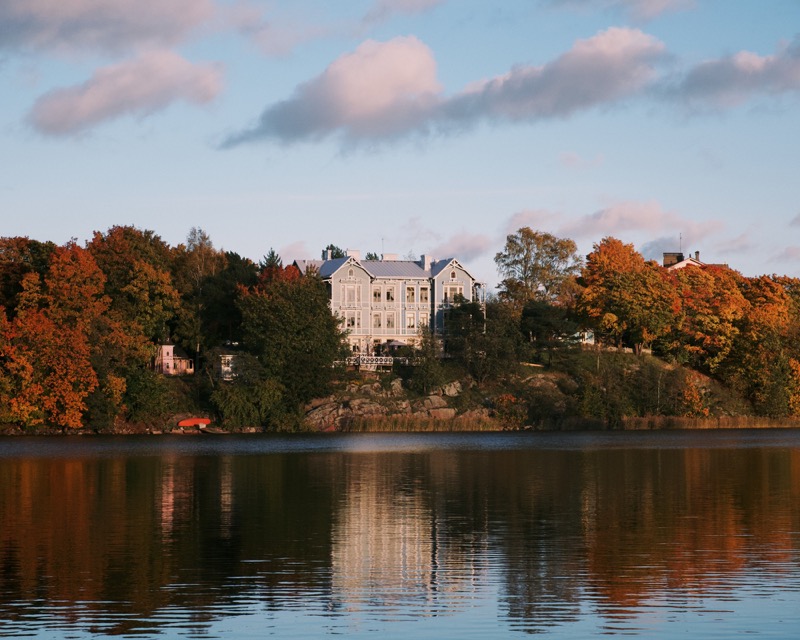 Large house across a lake, reflected in water beneath autumn trees.