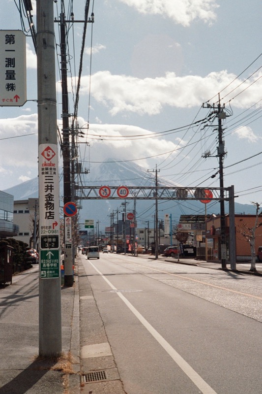 Quiet street lined with power lines and Japanese signs, with a mountain beyond.