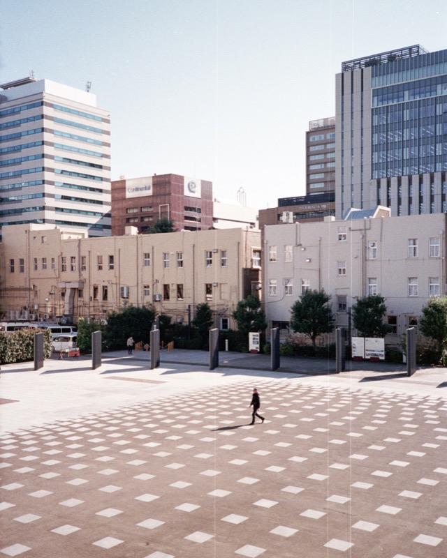 A lone pedestrian crosses a patterned plaza with office buildings in the background.