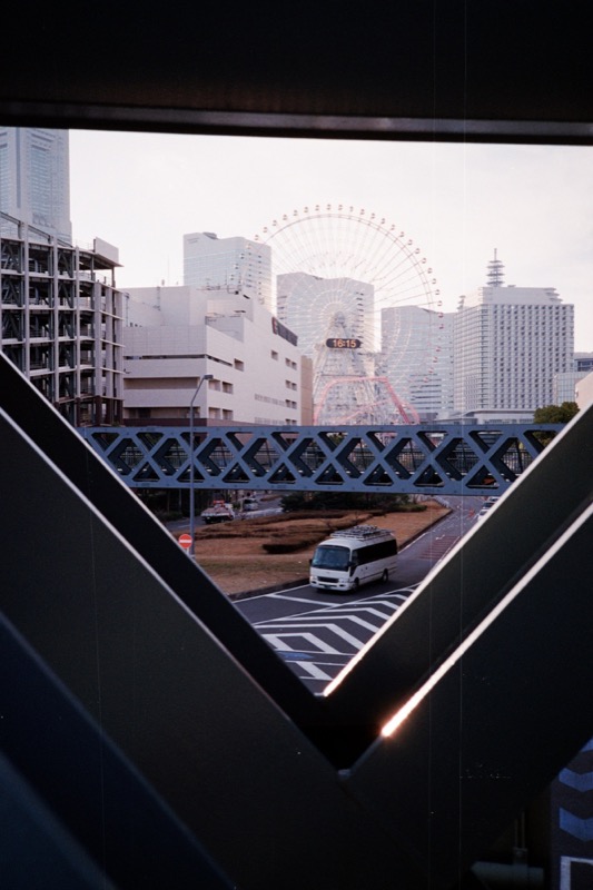 Ferris wheel and skyline seen through steel beams above a road with a bus.