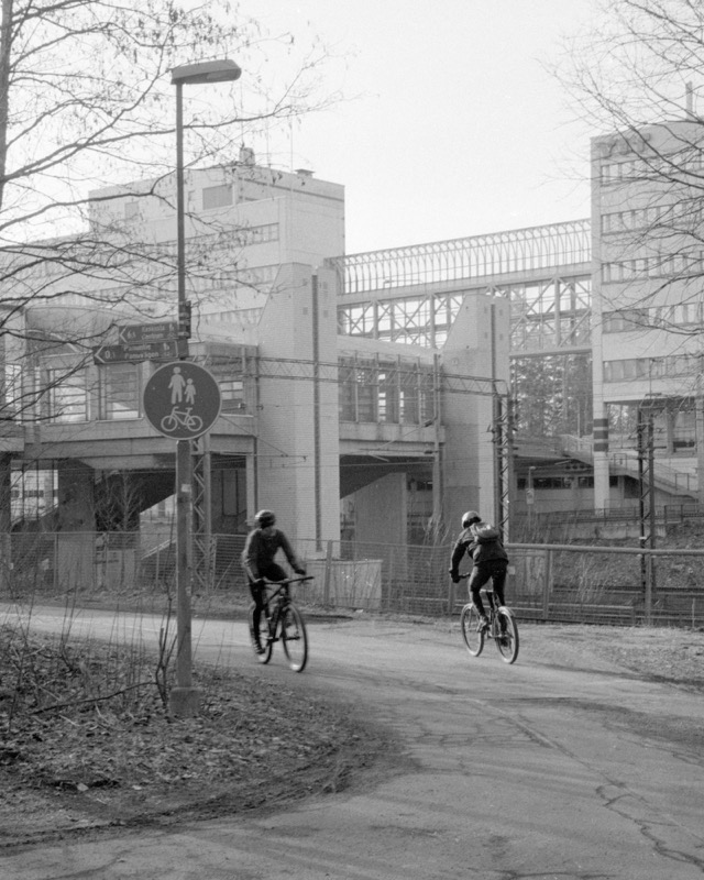 Two cyclists ride along a path near industrial buildings and elevated tracks.