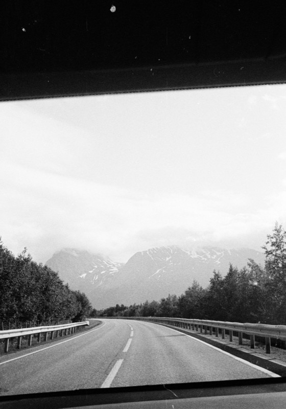 Empty highway curves toward snowy mountains, seen through a car windshield.