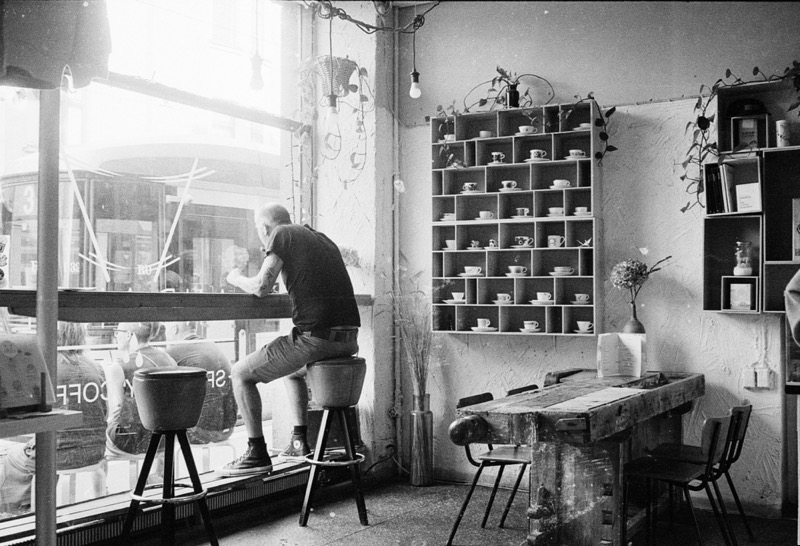 Man sits on a stool at a cafe window, beside shelves of cups.