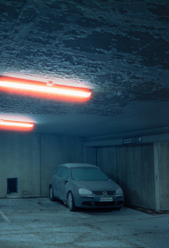 Dusty car sits in a dim concrete garage beneath glowing red lights.