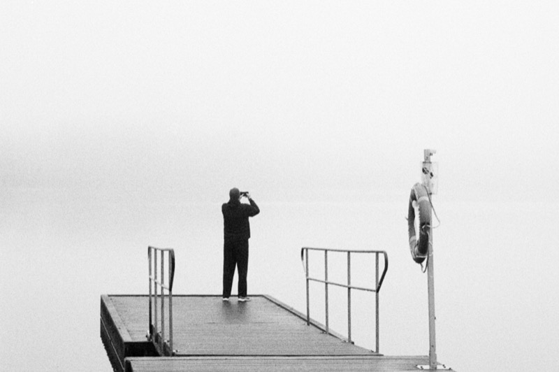 Solitary figure stands on a pier in fog, facing open water.