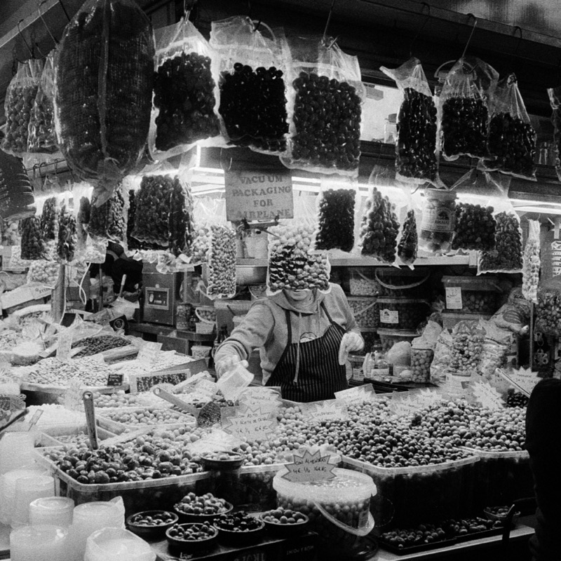 Market vendor reaches over bins of fruit beneath rows of hanging bagged produce.