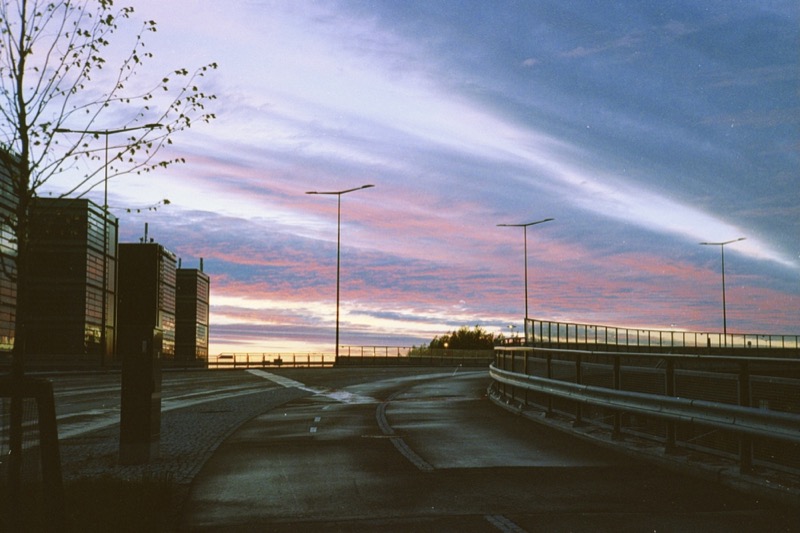 Empty elevated road curves past office buildings under a colorful sunset sky.