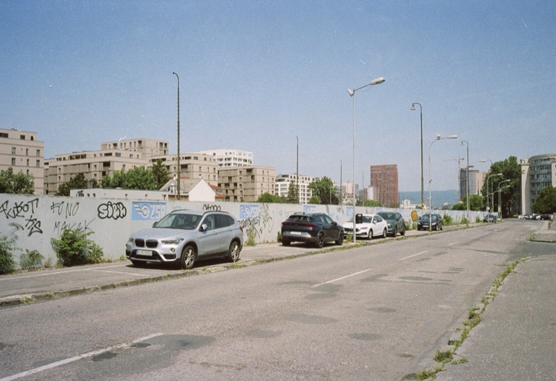 Parked cars line a quiet street beside a long graffiti wall in midday sun.