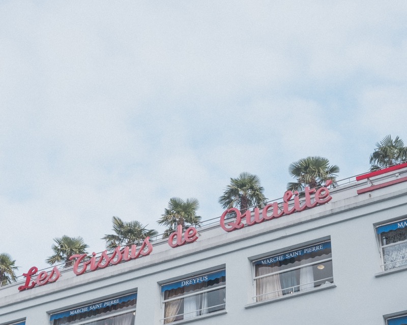 Rooftop sign reading 'Les Tissus de Qualité' above windows and palm trees.