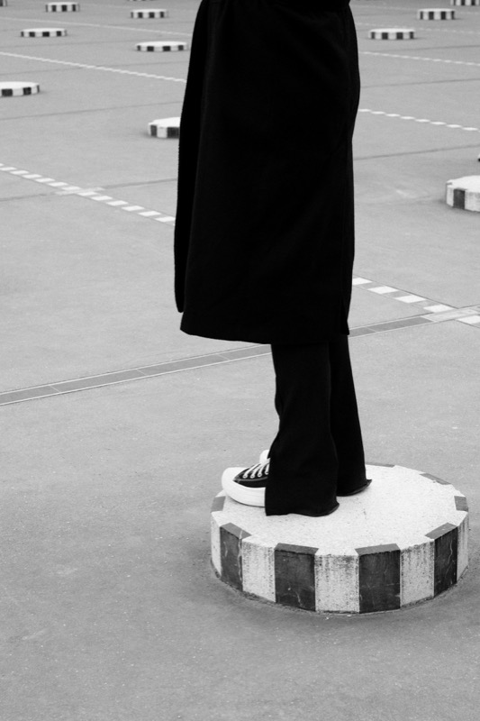Person in dark clothing stands on a round bollard in a paved lot.