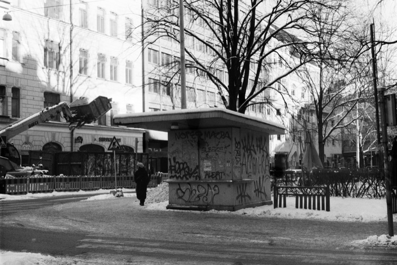 A graffitied kiosk on a snowy street, with a person walking past.