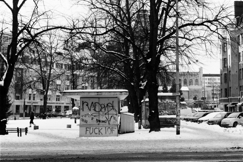 A kiosk wall covered in graffiti stands in a snowy lot beneath large bare trees.