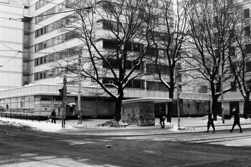 A long low kiosk along a modern building, with pedestrians crossing at a winter intersection.