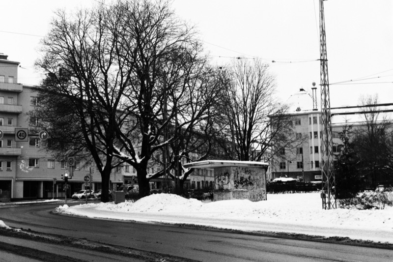 A graffitied kiosk sits in a snow-covered field beside a curving road and a large tree.