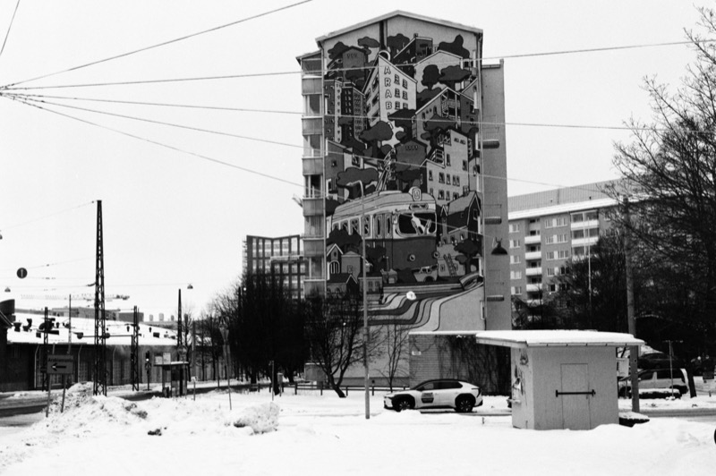 A small kiosk at the edge of a snowy lot, with a tall mural-covered apartment building behind.