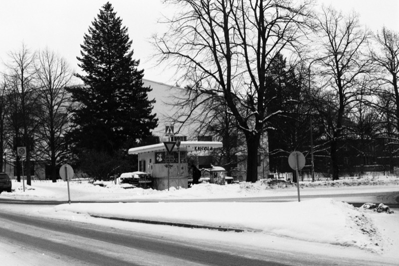 A shuttered kiosk at a snowy roadside, backed by a large pine tree and bare branches.