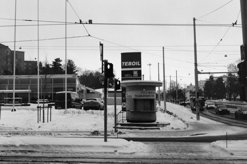 A small round kiosk at a snowy intersection beneath overhead wires and a Teboil sign.