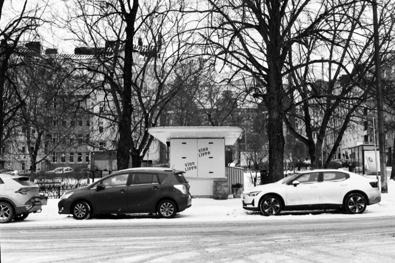 A boarded-up kiosk between parked cars on a snowy street, framed by tall bare trees.