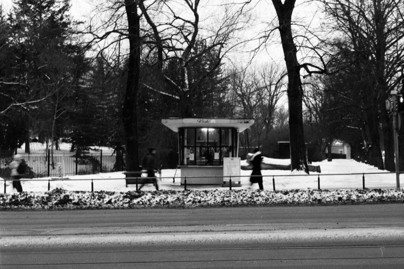 A kiosk with lit windows by the road, with motion-blurred pedestrians passing in the snow.