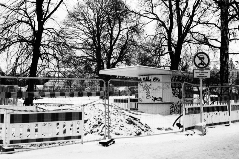 A graffitied kiosk behind temporary construction fencing in a snowy park.