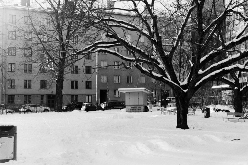 A small kiosk in a snow-covered courtyard framed by bare trees and an apartment block.