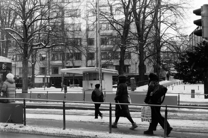Pedestrians cross a slushy street while a closed kiosk sits across the road in the snow.